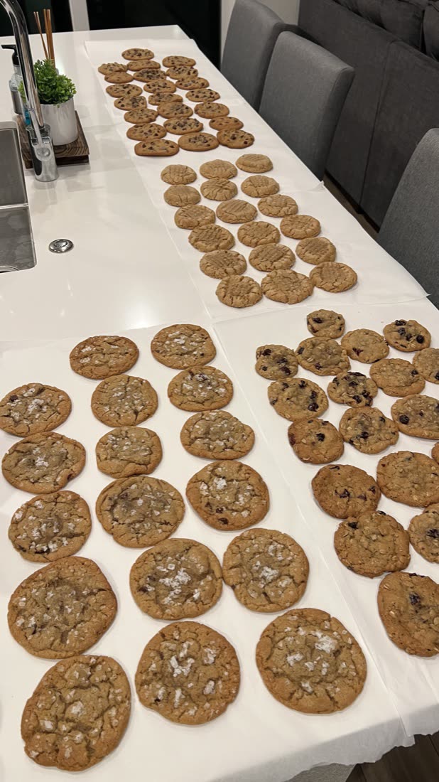 Dozens of cookies cooling in rows across a counter: chocolate chip, peanut butter, oatmeal cranberry, salted